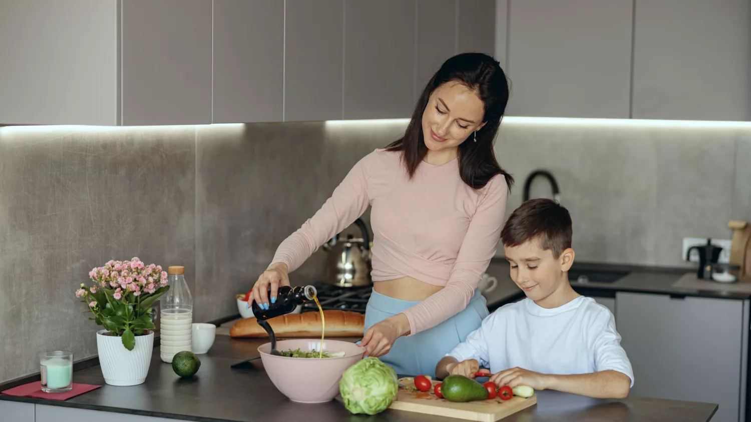 Mother cooking salad and putting olive oil her young son helping and chopping