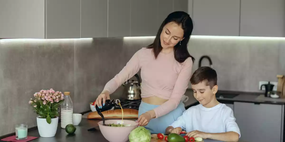 Mother cooking salad and putting olive oil her young son helping and chopping